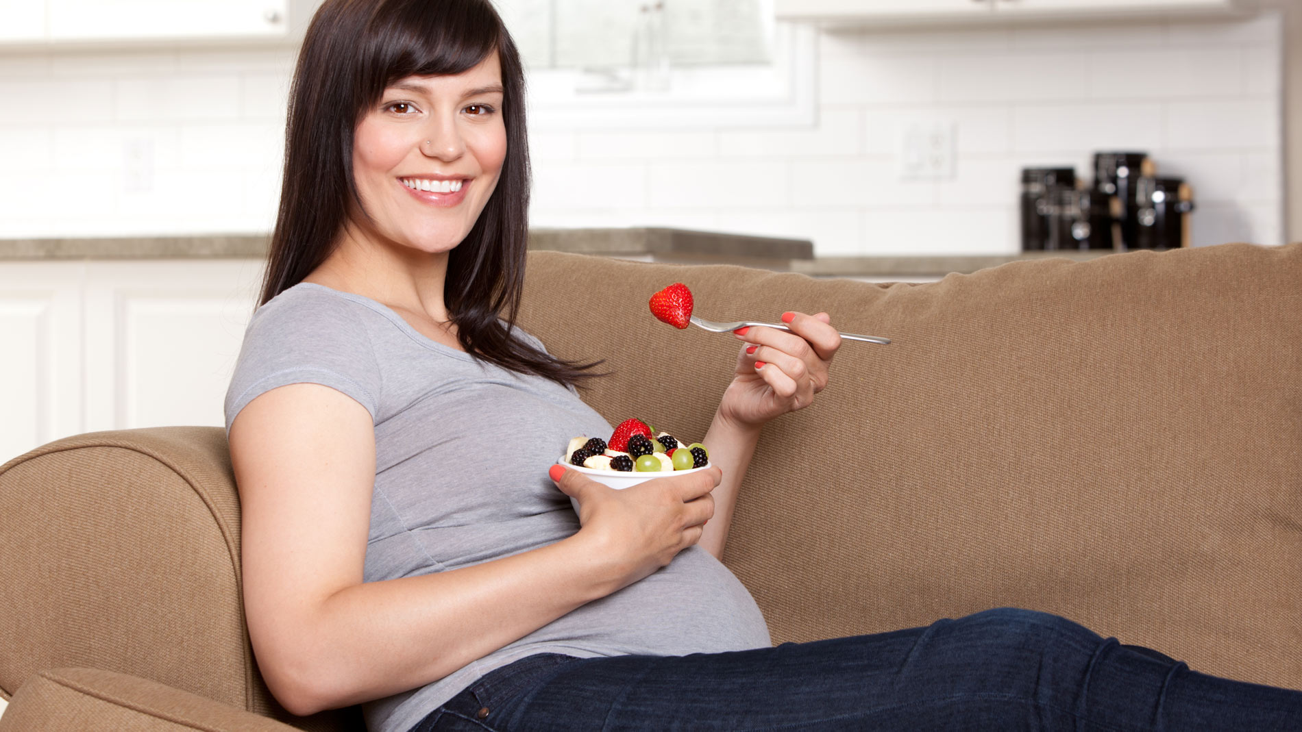 Happy pregnant woman sitting on a sofa eating bowl of fresh fruit