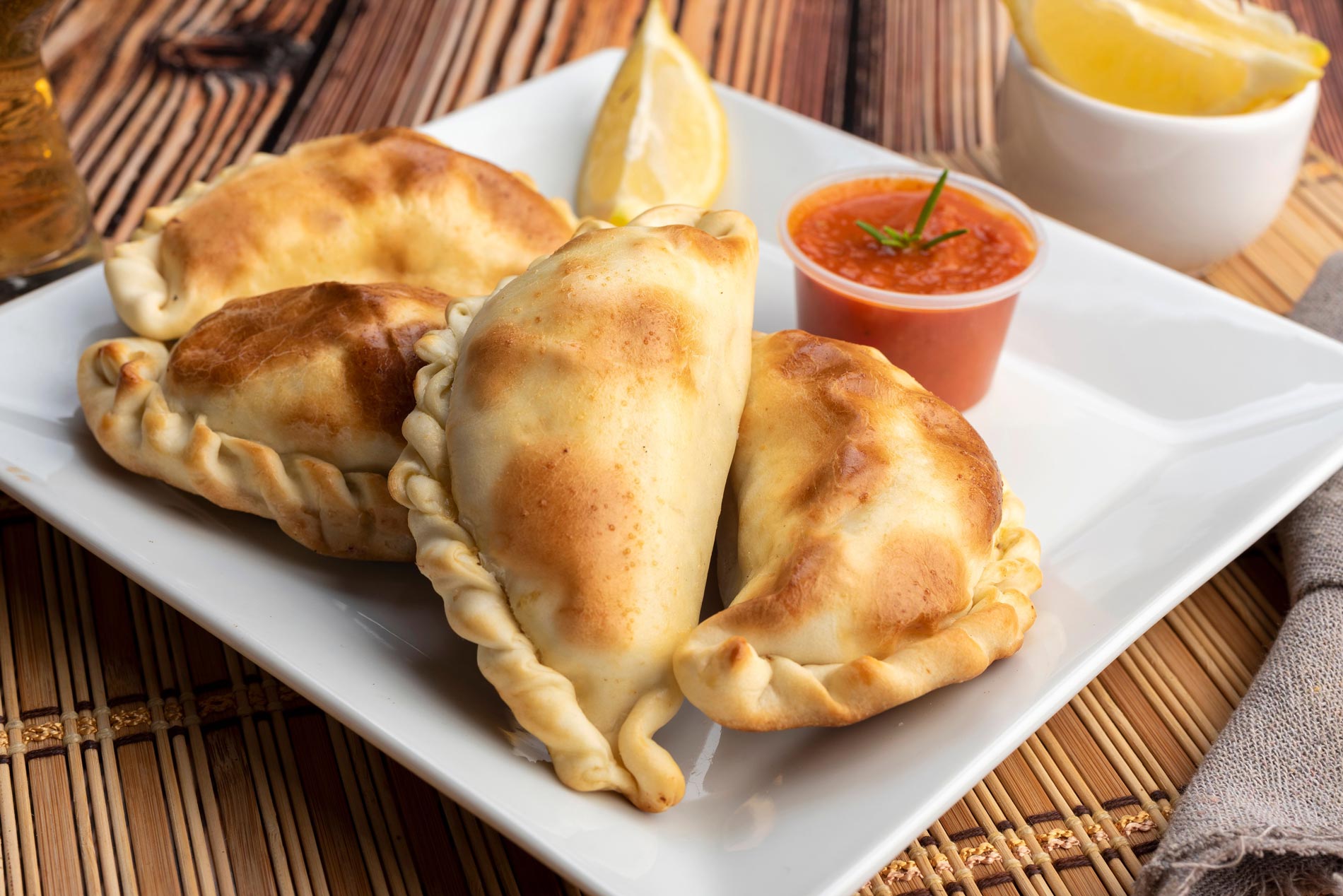 Argentinian homemade empanadas on square white plate, red sauce and lemon slices on wooden table