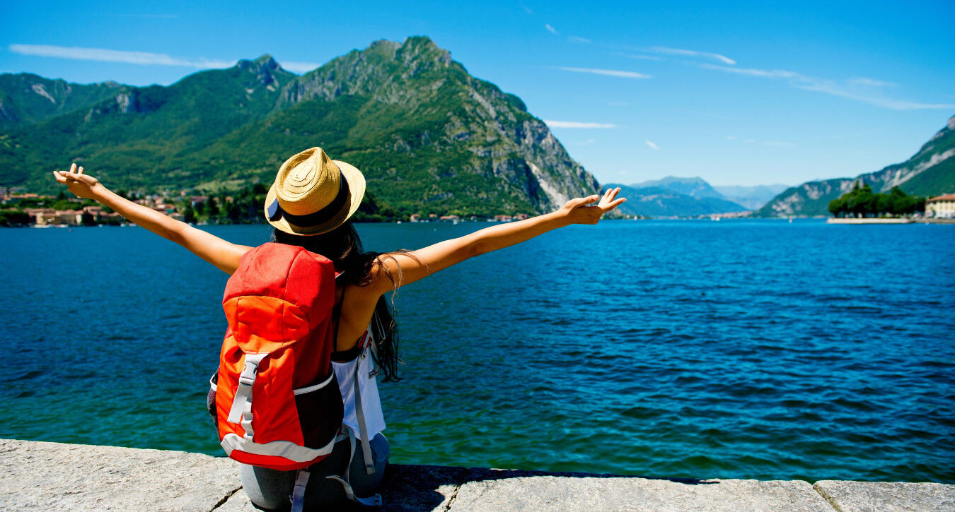 A traveller sits on a wall next to a mountainside lake with their arms open wide in appreciation