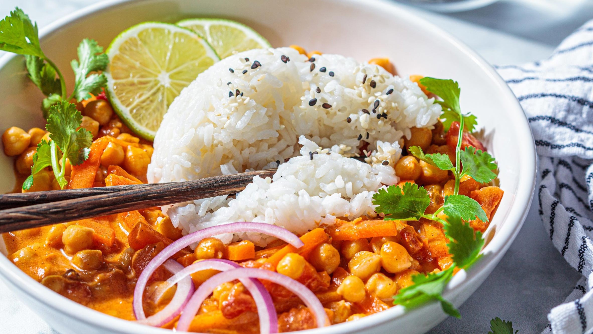 A bowl of white rice next to vegetables and chickpeas in red curry sauce and cilantro leaves for garnish