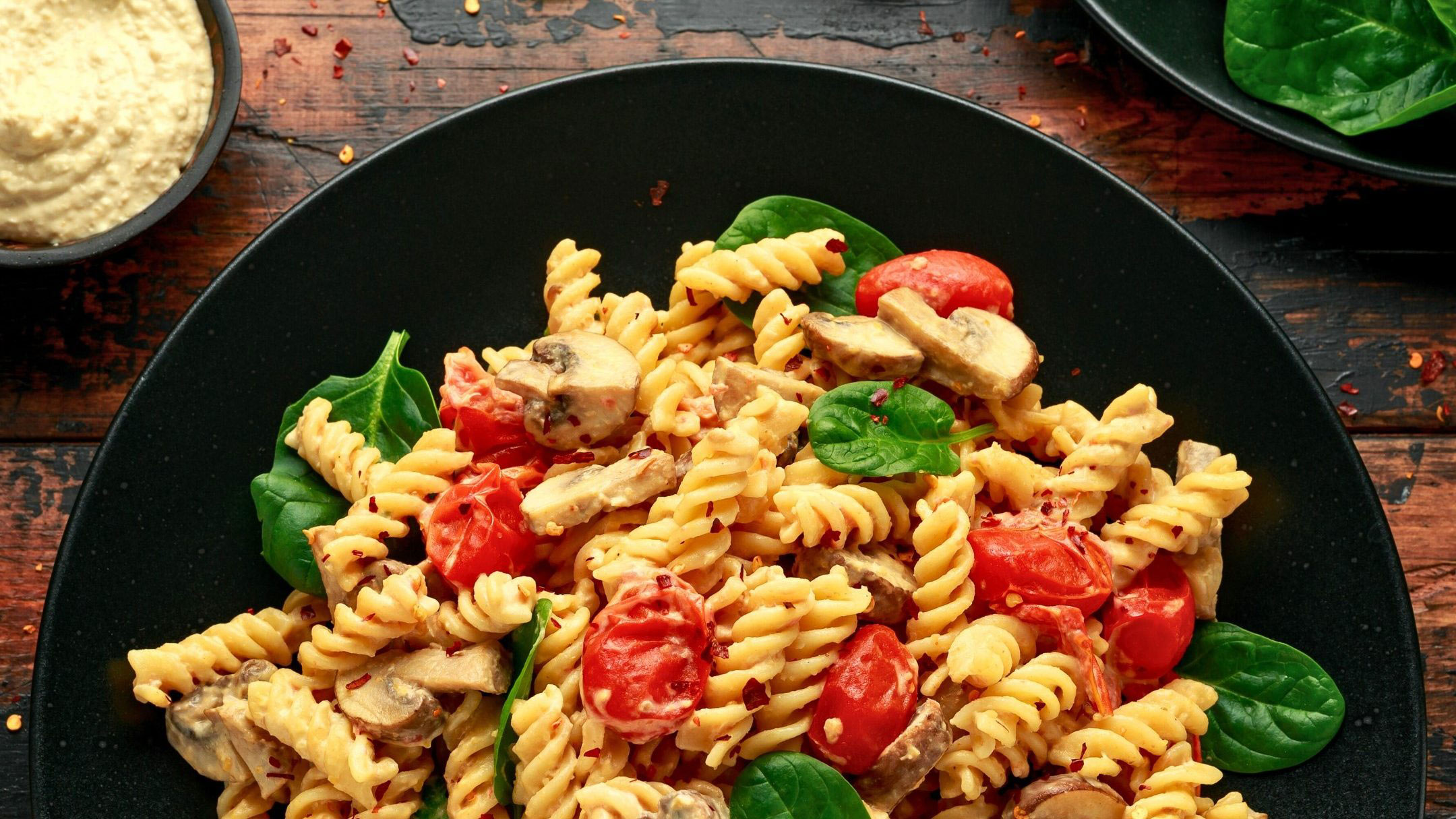 a bowl of cooked fusilli pasta in a thick creamy sauce with cherry tomatoes mushrooms and spinach leaves