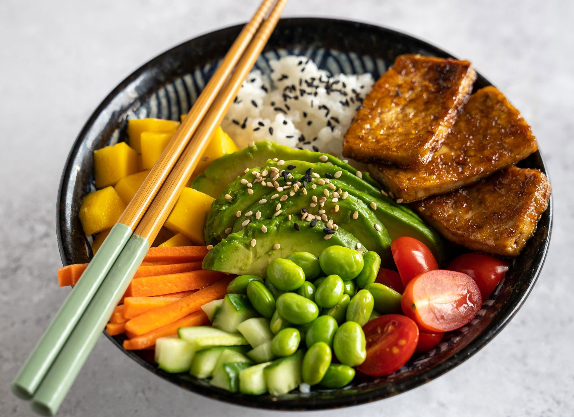 a bowl of rice, fresh vegetables, avocado, soy beans, and slices of tofu roasted with teriyaki